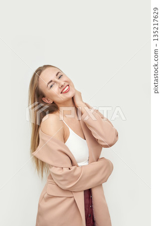 Young caucasian female smiling in beige coat with white top against light Young caucasian female smiling in beige coat with white top against light 135217629