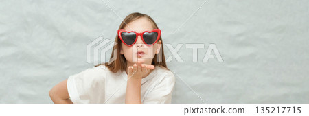 Caucasian young girl blowing kiss wearing red heart sunglasses against grey 135217715