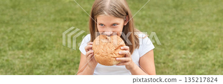 Caucasian young girl enjoying large bread loaf outdoors in green park setting 135217824