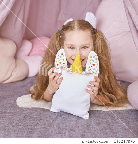 Happy caucasian female child with pigtails holding unicorn pillow in cozy pink 135217870