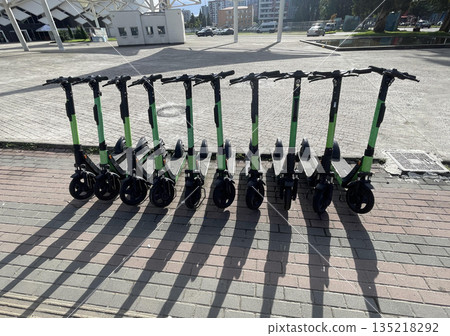 A row of scooters sits on the sidewalk for rent on a sunny day A row of scooters sits on the sidewalk for rent on a sunny day 135218292