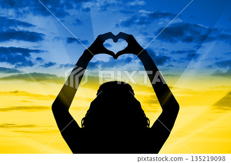 Silhouette of the woman's hands form a symbol heart on background of sky in the form of the flag of Ukraine. 135219098