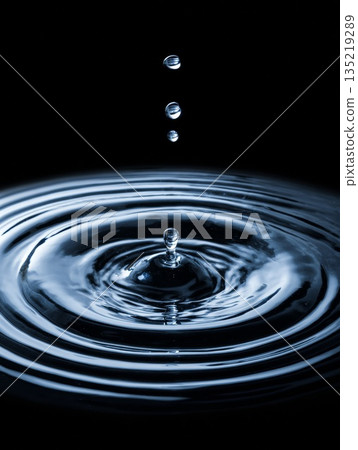 Water droplets falling into a calm surface creating ripples and reflections in dark background Water droplets falling into a calm surface creating ripples and reflections in dark background 135219289