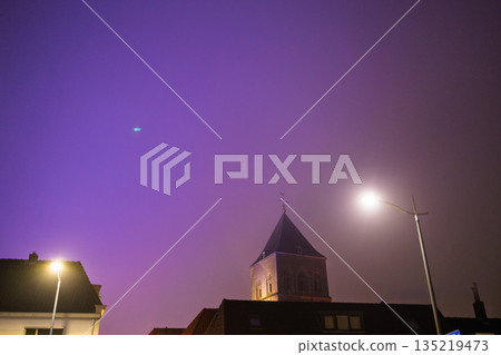 Nighttime Dutch town church tower lit beneath vivid purple sky, glowing streetlights and mysterious blue-green light evoke atmospheric intrigue. 135219473