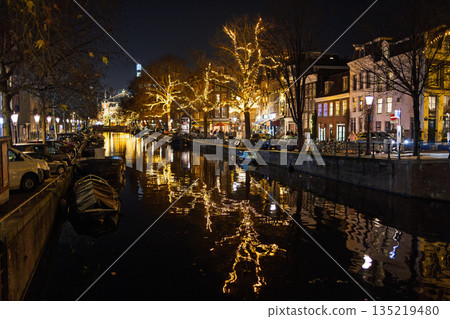 Amsterdam canal at night boats, festive lights on trees, glowing shopfronts, and reflections create a warm, vibrant winter scene. 135219480