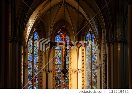 Interior of Gothic church central crucifix framed by vibrant stained glass, pointed arches, and ornate columns evoke reverence and spiritual grandeur. 135219481