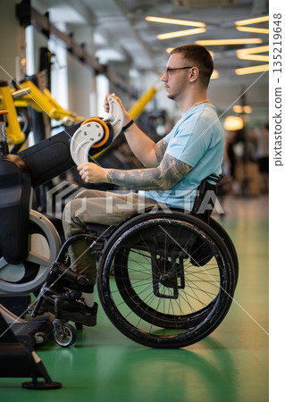Man in wheelchair works on upper body strength, turning levers on ergometer, do exercise in gym Man in wheelchair works on upper body strength, turning levers on ergometer, do exercise in gym 135219648