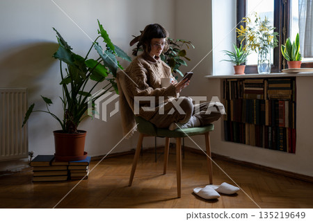 Teen girl sitting on chair with cup of tea, reading news, scrolling photos, messaging in smartphone 135219649