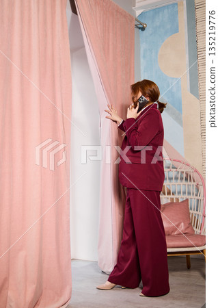 Woman in maroon outfit checks through curtain calmly during call Woman in maroon outfit checks through curtain calmly during call 135219776