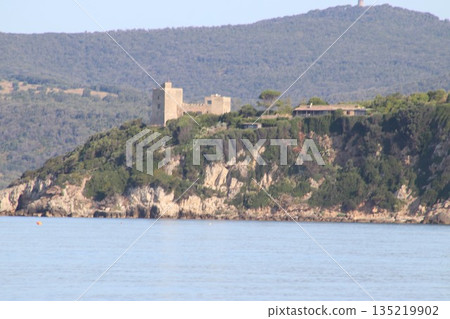 A close-up view of the historic Aldobrandeschi Castle situated on a rugged promontory surrounded by lush Mediterranean vegetation and calm blue water in the Maremma park area. 135219902