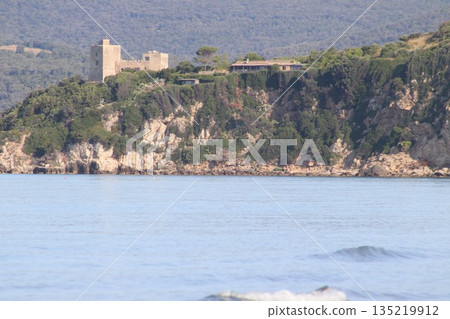 A close-up view of the historic Aldobrandeschi Castle situated on a rugged promontory surrounded by lush Mediterranean vegetation and calm blue water in the Maremma park area. A close-up view of the historic Aldobrandeschi Castle situated on a rugged promontory surrounded by lush Mediterranean vegetation and calm blue water in the Maremma park area. 135219912