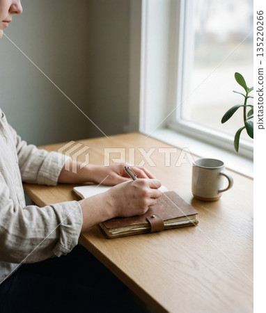 Person writing in journal at wooden desk by window Person writing in journal at wooden desk by window 135220266