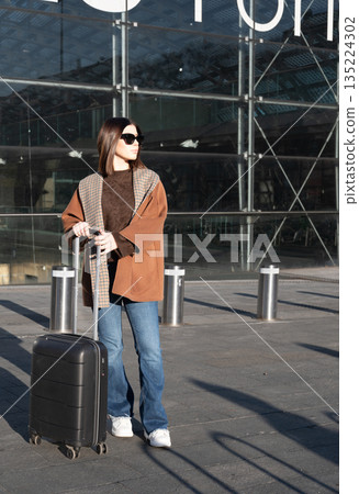 Woman wearing sunglasses and coat, pulling her black suitcase, walking outdoors near a modern glass building, representing travel, business trips, and vacation 135224302