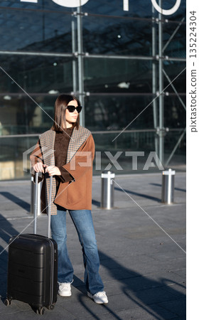 Woman standing outdoors at an airport or terminal entrance with a roller suitcase, wearing sunglasses, a brown coat, and jeans, preparing for a journey Woman standing outdoors at an airport or terminal entrance with a roller suitcase, wearing sunglasses, a brown coat, and jeans, preparing for a journey 135224304