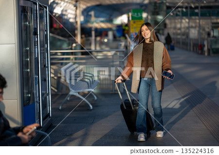 Young woman pulling a suitcase along a busy train platform, smiling and holding a smartphone as she prepares to board and continue her urban journey and exploration Young woman pulling a suitcase along a busy train platform, smiling and holding a smartphone as she prepares to board and continue her urban journey and exploration 135224316