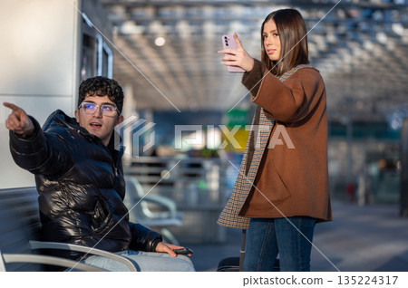 Young couple traveling in a modern train station, the man pointing while sitting and the woman standing holding a mobile phone looking for information or taking a selfie Young couple traveling in a modern train station, the man pointing while sitting and the woman standing holding a mobile phone looking for information or taking a selfie 135224317