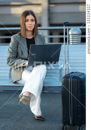 Young businesswoman sits beside her suitcase on a bench, typing on a laptop in a city waiting area, embodying remote work, digital nomad lifestyle and business travel connectivity 135224327