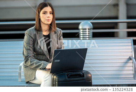 Female entrepreneur sitting on a bench with her suitcase, focused on her laptop, embodying remote work and business travel freedom in a modern setting 135224328