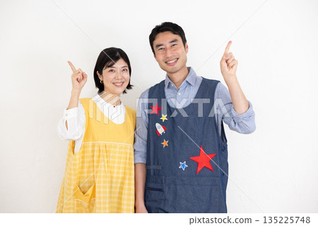 Smiling male and female nursery teachers wearing aprons smiling with their index fingers raised Smiling male and female nursery teachers wearing aprons smiling with their index fingers raised 135225748
