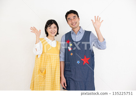 Smiling male and female nursery teachers wearing aprons waving with a smile Smiling male and female nursery teachers wearing aprons waving with a smile 135225751