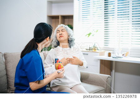 Asian nurse explains a heart model to an elderly woman at home, focusing on heart health, cardiology, patient education, senior care, and modern home 135226978