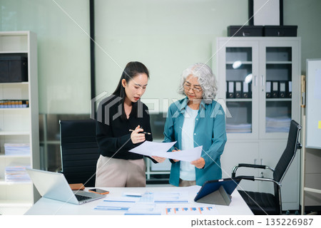 Asian senior and middle aged businesswomen reviewing documents and reports in office. Professional teamwork, mentorship, business collaboration Asian senior and middle aged businesswomen reviewing documents and reports in office. Professional teamwork, mentorship, business collaboration 135226987