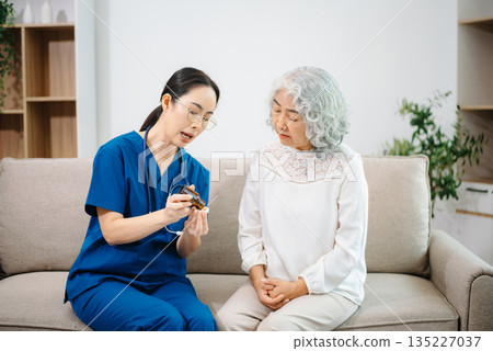 Young nurse holding a bottle of pills and discussing treatment with an elderly Asian woman during a medical consultation Young nurse holding a bottle of pills and discussing treatment with an elderly Asian woman during a medical consultation 135227037