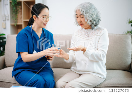 Young nurse holding a bottle of pills and discussing treatment with an elderly Asian woman during a medical consultation Young nurse holding a bottle of pills and discussing treatment with an elderly Asian woman during a medical consultation 135227073