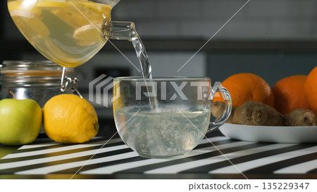 Hand pouring hot lemon ginger tea into a glass cup. Soothing drink for wellness and home comfort concept Hand pouring hot lemon ginger tea into a glass cup. Soothing drink for wellness and home comfort concept 135229347