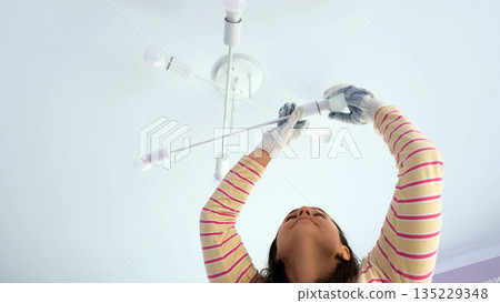 A woman stands on a stepladder carefully screwing in a light bulb into a ceiling lamp wearing gloves and focused on her task in a bright interior space A woman stands on a stepladder carefully screwing in a light bulb into a ceiling lamp wearing gloves and focused on her task in a bright interior space 135229348