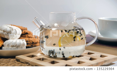 Hot water poured into glass teapot with green loose leaf tea and lemon, with cookies adding to this relaxing tea experience  135229364