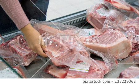 Close-up of woman choosing packaged pork in a supermarket freezer, checking label for price and freshness 135229368