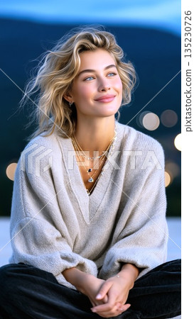 Blonde woman smiling, wearing beige sweater and gold necklaces, sitting cross legged against soft urban sunset background with blue eyed gaze,Blonde woman 135230726