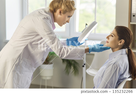 Dermatologist inspecting patient face with magnifying lamp in modern clinic Dermatologist inspecting patient face with magnifying lamp in modern clinic 135232266