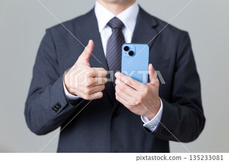 A middle-aged male businessman in a suit holding a smartphone and giving a thumbs up A middle-aged male businessman in a suit holding a smartphone and giving a thumbs up 135233081