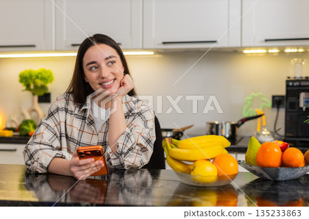 Young woman chats on smartphone at kitchen table scrolling news and buy online alone indoors at home Young woman chats on smartphone at kitchen table scrolling news and buy online alone indoors at home 135233863