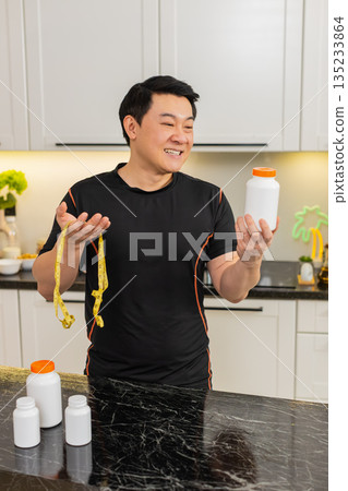 Asian man in kitchen holding supplement bottle with tape measure celebrating fit healthy weight loss Asian man in kitchen holding supplement bottle with tape measure celebrating fit healthy weight loss 135233864