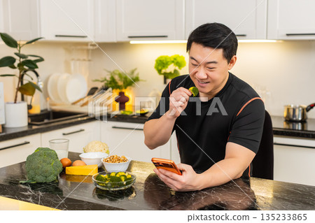 Asian man eats broccoli near smartphone in kitchen while reading health news and planning clean diet Asian man eats broccoli near smartphone in kitchen while reading health news and planning clean diet 135233865