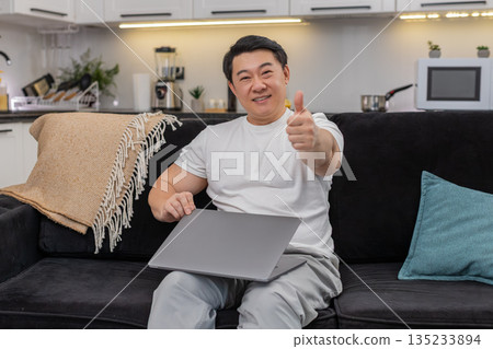 Asian man closes laptop after tasks exhales stretches smiles pride shows big thumbs up toward camera 135233894