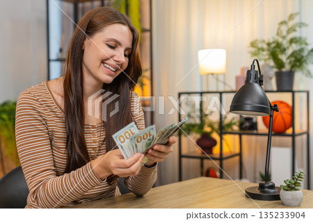 Woman counts cash dollar bills money at table smiles pleased about salary lottery win savings income Woman counts cash dollar bills money at table smiles pleased about salary lottery win savings income 135233904