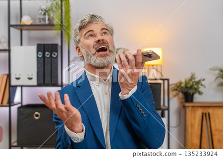 Middle-aged businessman argues on speakerphone smartphone angry gestures about shipping delay issues Middle-aged businessman argues on speakerphone smartphone angry gestures about shipping delay issues 135233924