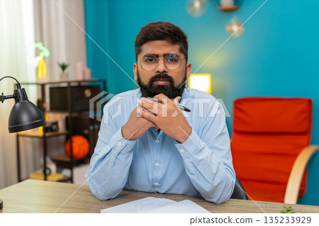 Indian man holds pen gazes notebook deeply thinking planning ideas writing strategy indoors focused 135233929