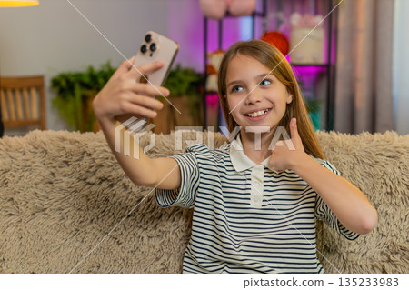 Child girl kid happily takes selfie on smartphone makes funny faces and smiles while sitting on sofa Child girl kid happily takes selfie on smartphone makes funny faces and smiles while sitting on sofa 135233983