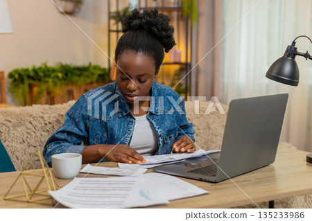 African American young woman checks utility bills on laptop sitting at home table with documents 135233986