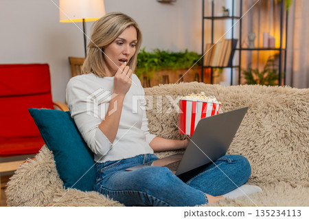 Adult woman stunned by shocking movie moment at home, holding popcorn and staring at laptop screen 135234113