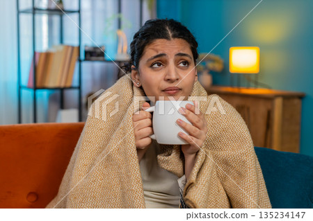 Indian woman on sofa wrapped in blanket holding hot tea cup feeling cold autumn winter weather 135234147