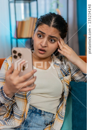 Indian woman sadly looking at smartphone on sofa disappointed receiving bad news breakup or losing 135234148