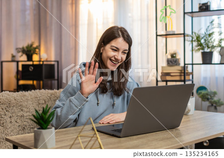 Happy young woman sitting at table talking to friend by video call using laptop wave hand at home Happy young woman sitting at table talking to friend by video call using laptop wave hand at home 135234165