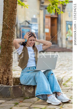 Caucasian young woman using laptop browsing loses lottery results, bad news sitting in city street Caucasian young woman using laptop browsing loses lottery results, bad news sitting in city street 135234211