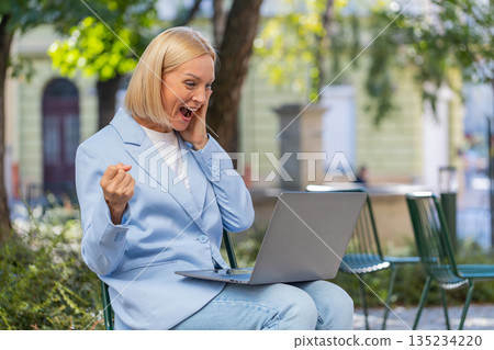 Happy mature businesswoman using laptop computer celebrating success victory clenching fists in city 135234220